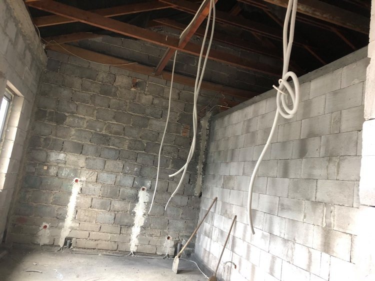 Wnętrze surowego pomieszczenia z widocznymi przewodami elektrycznymi wystającymi ze ściany z betonu komórkowego, przygotowane otwory pod gniazdka, surowe drewniane belki stropowe.