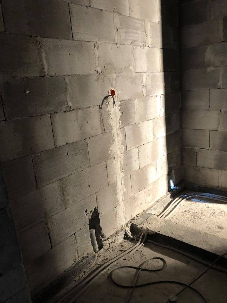 Surowa ściana z betonu komórkowego z widoczną puszką elektryczną z wystającymi przewodami oraz peszlami na podłodze w trakcie prac instalacyjnych.