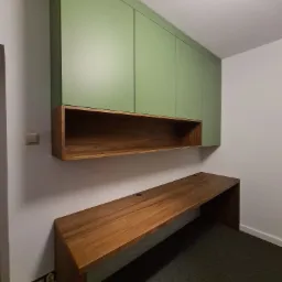 Zestaw mebli: wiszące szafki z zielonymi frontami nad drewnianą półką i długim blatem biurka z tego samego drewna, na tle białej ściany i szarej wykładziny.