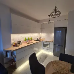 Nowoczesna kuchnia z białymi frontami, drewnianym blatem, marmurowym backsplash'em i oświetleniem LED pod szafkami, widoczny stół z plecionymi podkładkami i dekoracyjne lampy wiszące.