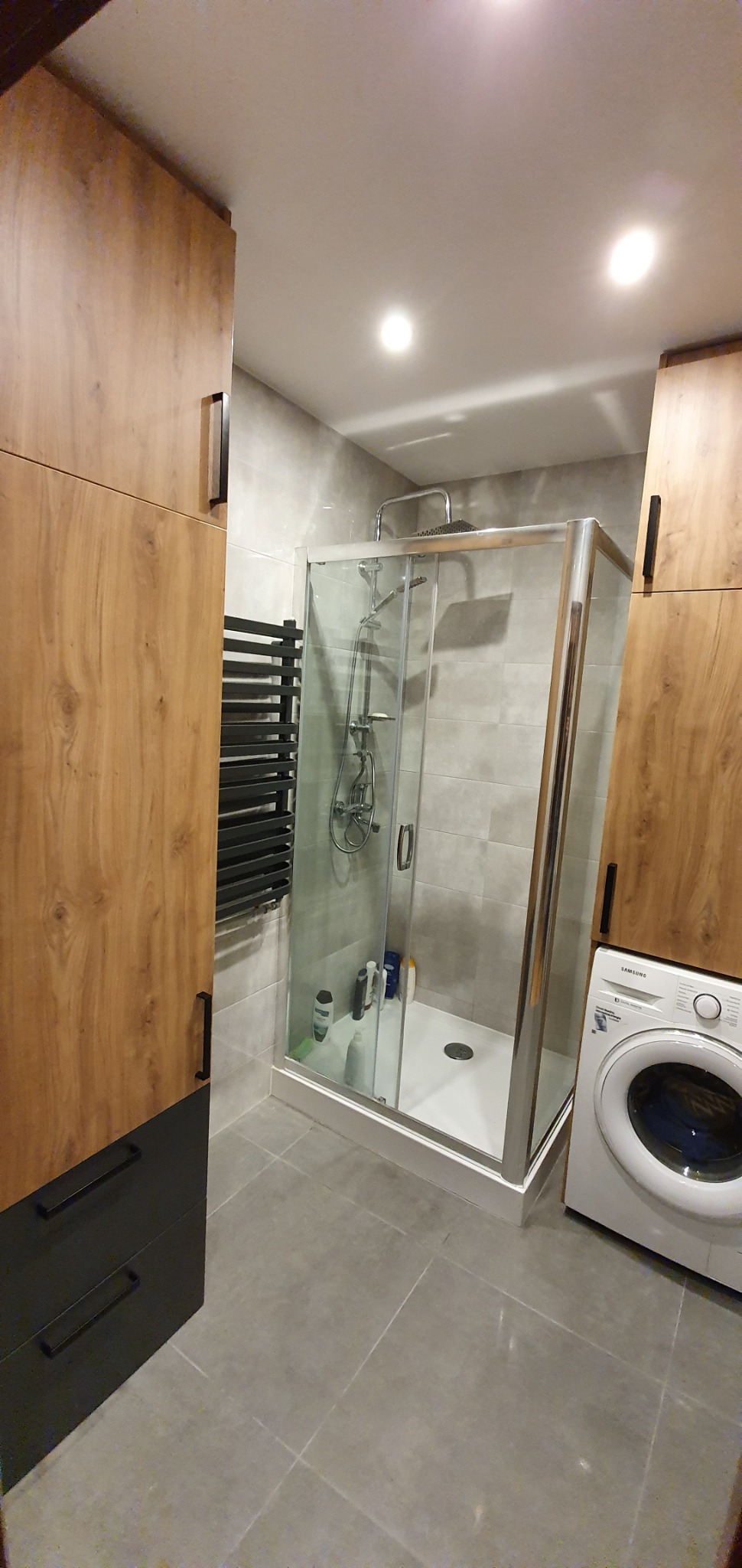 Nowoczesna łazienka z kabiną prysznicową, grzejnikiem drabinkowym i szafkami z drewnopodobnym wykończeniem oraz pralką. Szare płytki i oświetlenie punktowe.