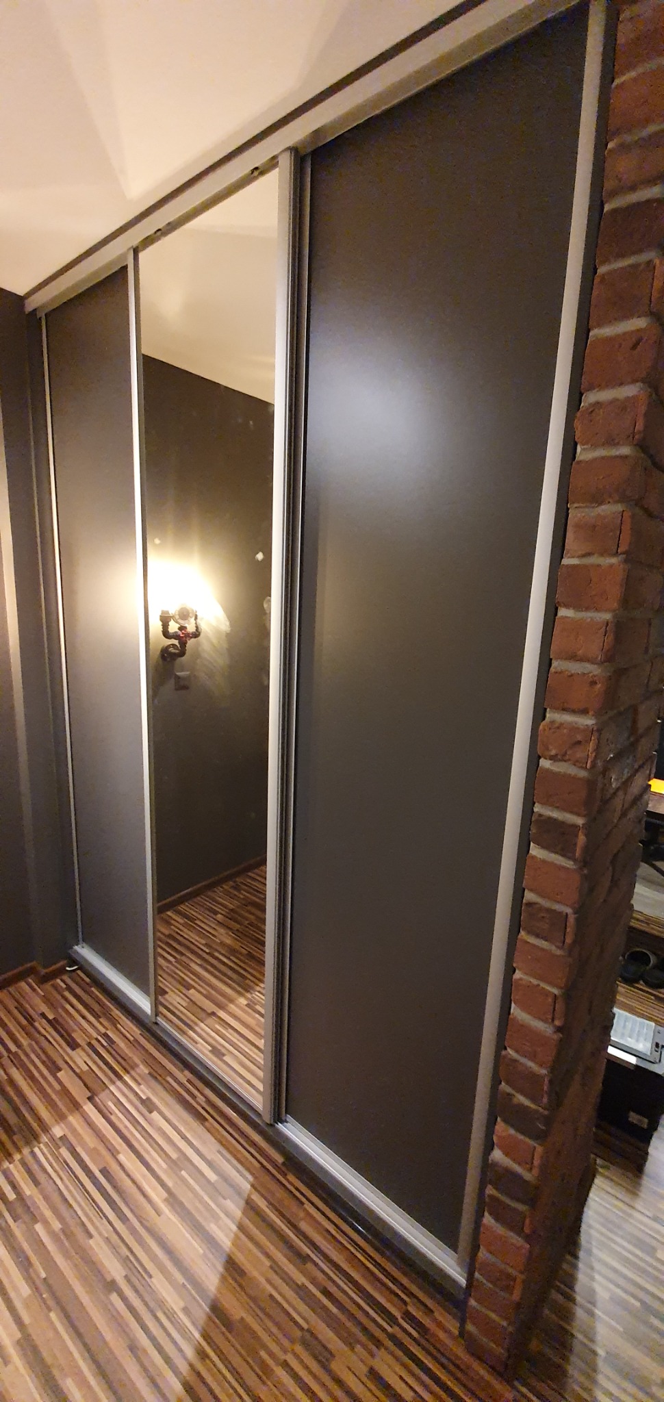 Nowoczesna szafa przesuwna z lustrem, wkomponowana w przestrzeń z ceglaną ścianą i drewnianą podłogą, oświetlona lampą industrialną.