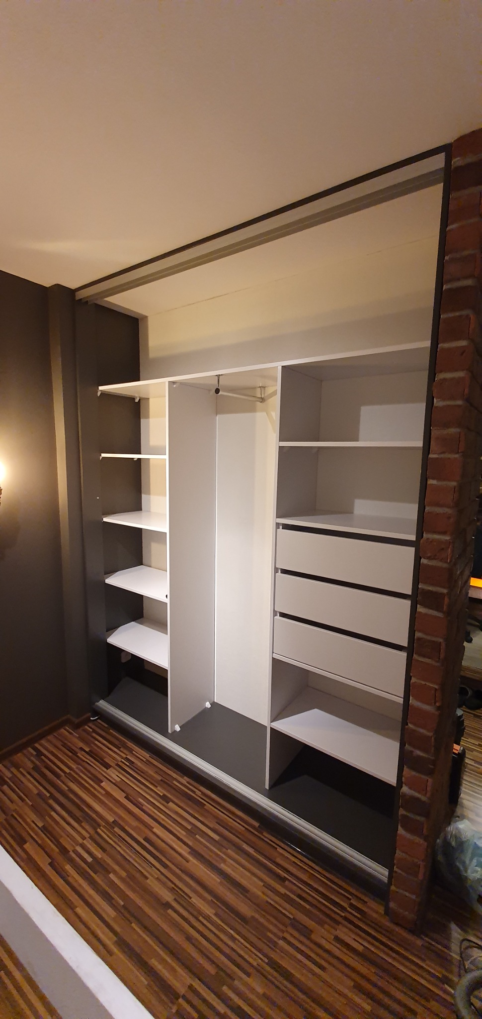 Wnętrze szafy wnękowej z białymi półkami i szufladami, zamontowanej między ciemnoszarą ścianą a ścianą z czerwonej cegły, widoczny fragment drewnianej podłogi.