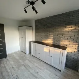 Nowoczesne wnętrze z białą komodą i szafą na tle ściany z szarej cegły, oświetlone reflektorami sufitowymi, z widoczną podłogą imitującą drewno.