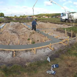 Wylewanie betonowych fundamentów pod budynek jednorodzinny; widoczne szalunki, zbrojenia, pracownik w niebieskiej bluzie oraz dwa betonomieszarki w tle.