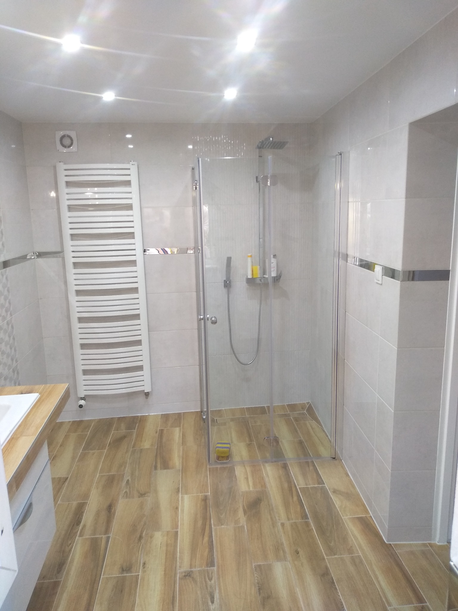 Wykończona łazienka z kabiną prysznicową typu walk-in, grzejnikiem drabinkowym i płytkami imitującymi drewno na podłodze.