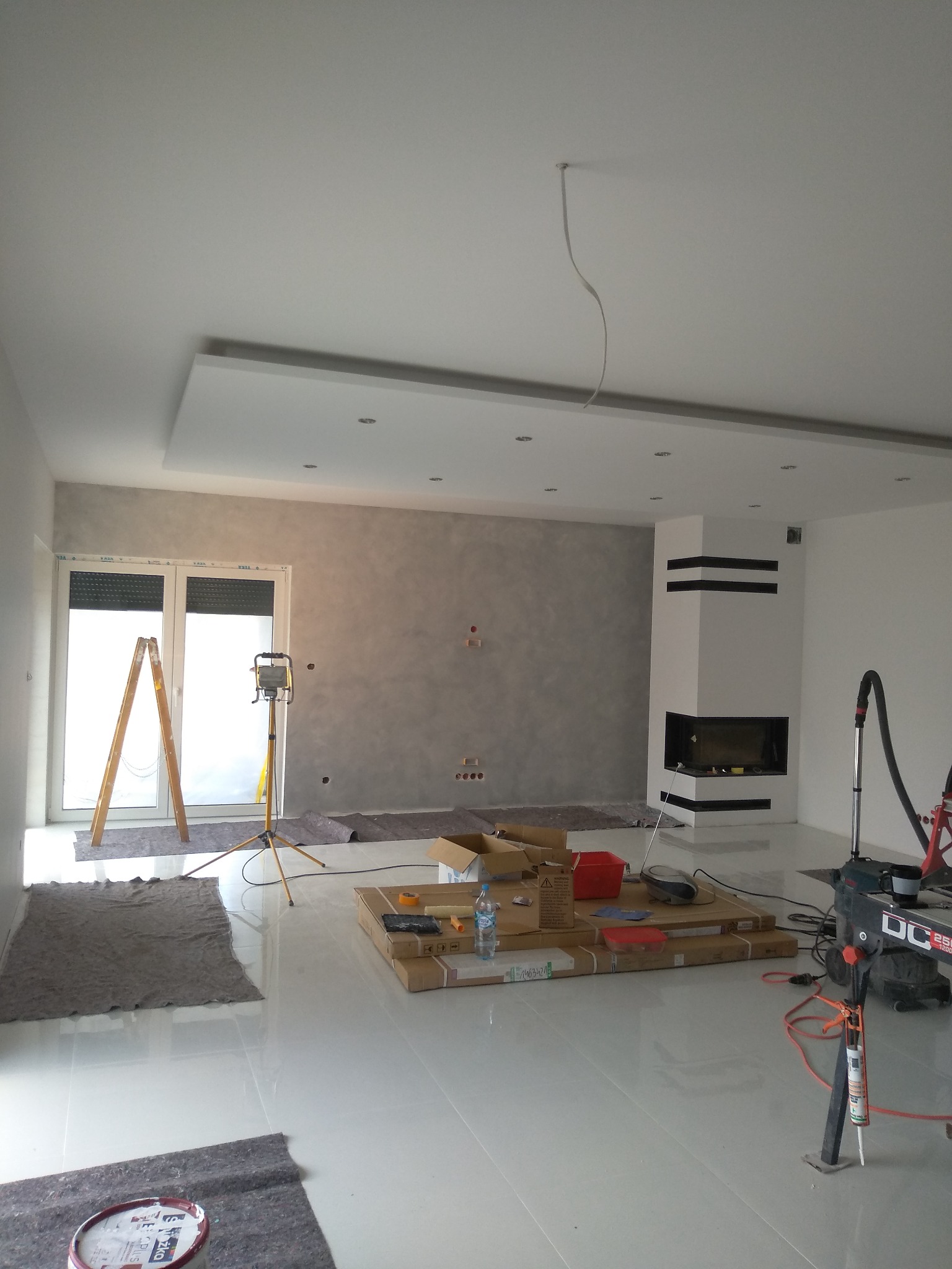 Wnętrze w trakcie wykańczania: ściana z dekoracyjnym tynkiem, biały kominek z czarnymi akcentami, rozstawiona drabina i lampa budowlana, zabezpieczona podłoga, kartony z materiałami i narzędzia.