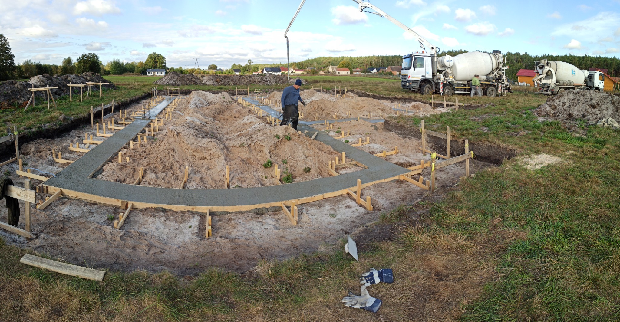 Wylewanie betonowych fundamentów pod budynek jednorodzinny; widoczne szalunki, zbrojenia, pracownik w niebieskiej bluzie oraz dwa betonomieszarki w tle.