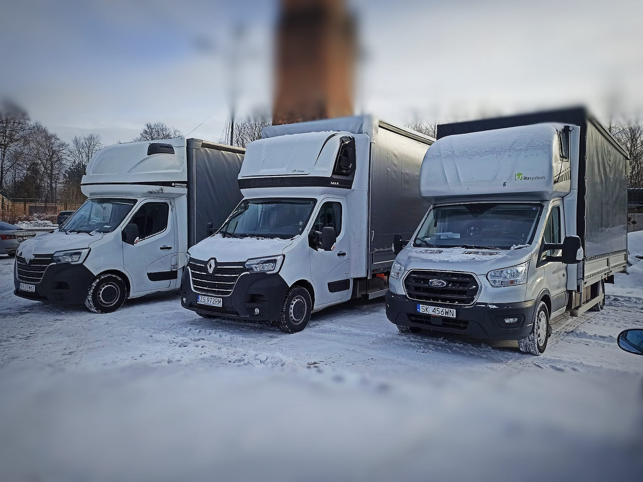 Trzy białe busy dostawcze różnych marek (Renault, Ford i Partner) zaparkowane obok siebie na śniegu, każdy z zabudową kontenerową, widoczne tablice rejestracyjne.