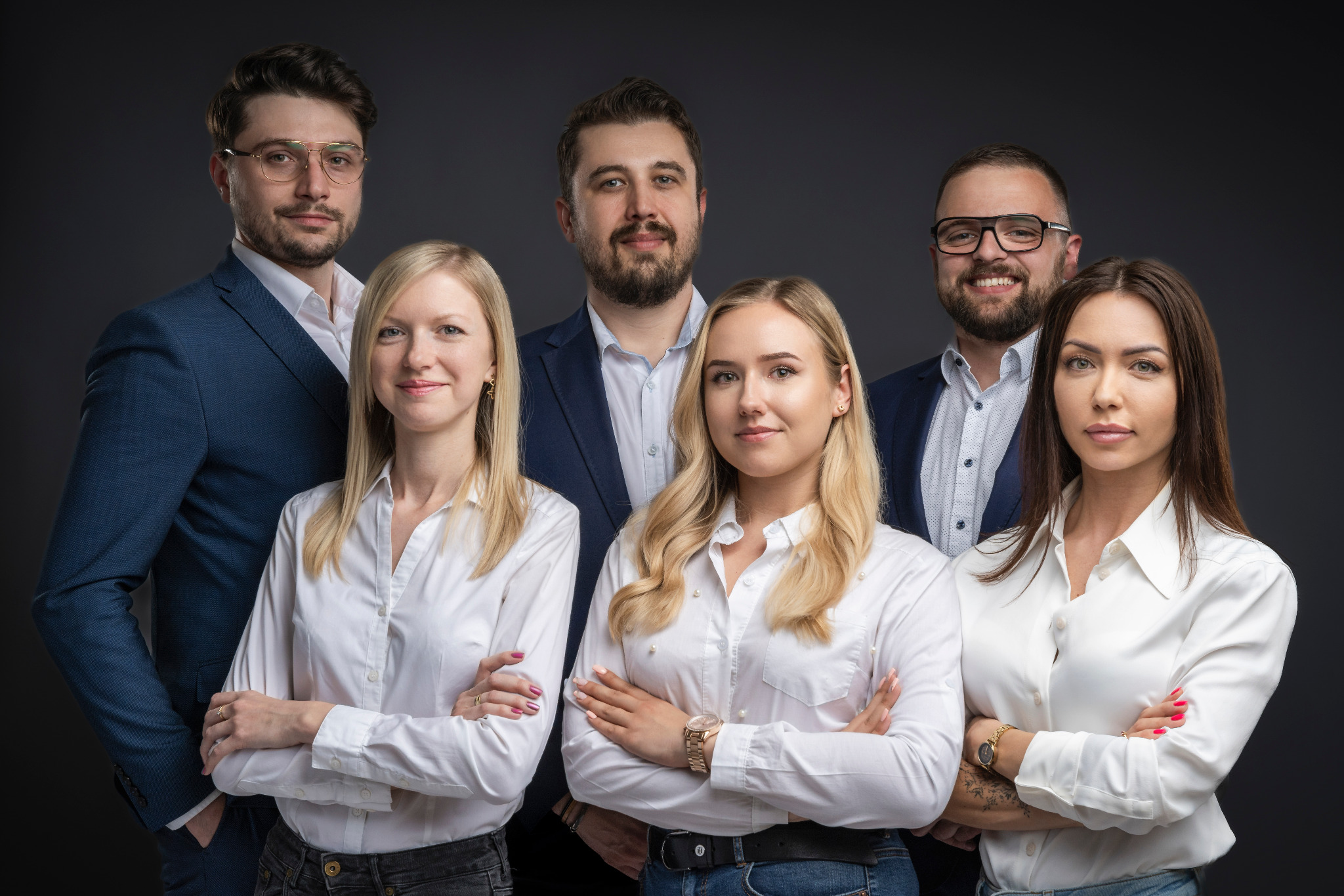 Zespół sześciu osób, trzy kobiety i trzech mężczyzn, w formalnych strojach pozuje z założonymi rękami na ciemnoszarym tle, sugerując profesjonalizm i zaufanie.