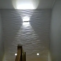Klatka schodowa z drewnianymi schodami i balustradą, ściana wykończona panelami 3D w kształcie fal, oświetlenie punktowe na ścianie.