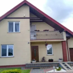 Dwupiętrowy dom z beżową elewacją, czerwonym dachem i balkonem z kutą balustradą, otoczony zielonym trawnikiem i dekoracyjnymi roślinami w donicach.