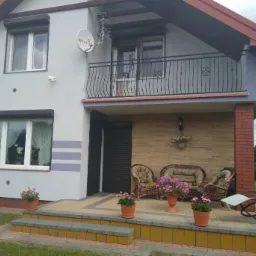 Dwupiętrowy dom z szarą elewacją, roletami zewnętrznymi, balkonem z czarną balustradą i tarasem z rattanowymi meblami i kwiatami w doniczkach.