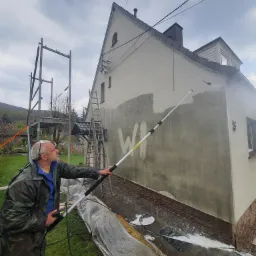 Mężczyzna w kombinezonie ochronnym myje elewację domu za pomocą lancy ciśnieniowej, widoczne rusztowanie i drabina oparta o ścianę.
