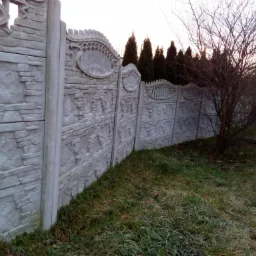 Długi, szary, betonowy płot z dekoracyjnym wzorem, widziany pod kątem, z drzewami iglastymi w tle.