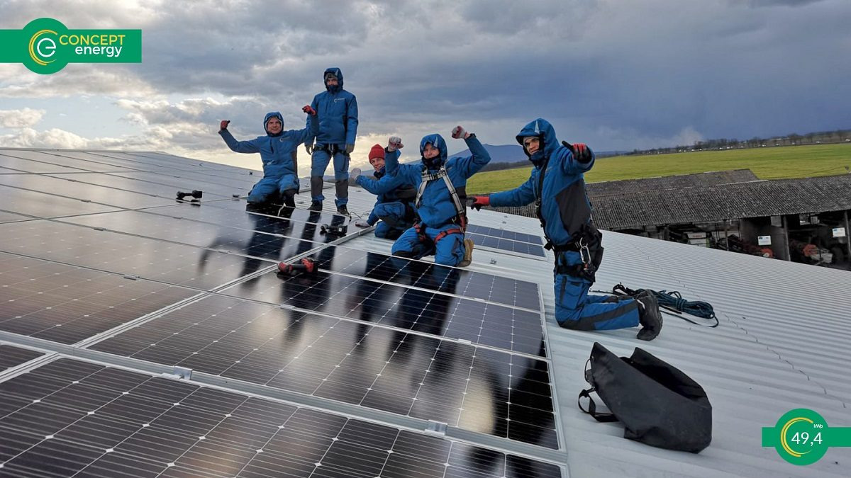 Grupa monterów w niebieskich kombinezonach świętuje ukończenie instalacji paneli słonecznych na dachu budynku gospodarczego, widok na zielone pola i pochmurne niebo w tle.