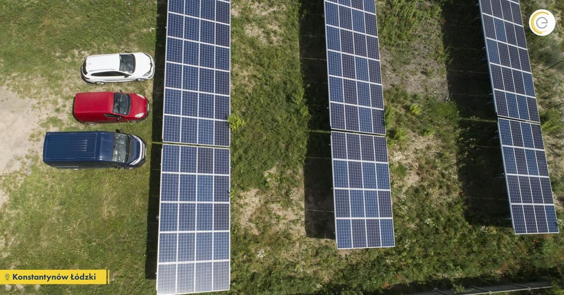 Widok z góry na rząd paneli słonecznych obok zaparkowanych samochodów: białego, czerwonego i niebieskiego vana na tle zielonej trawy.