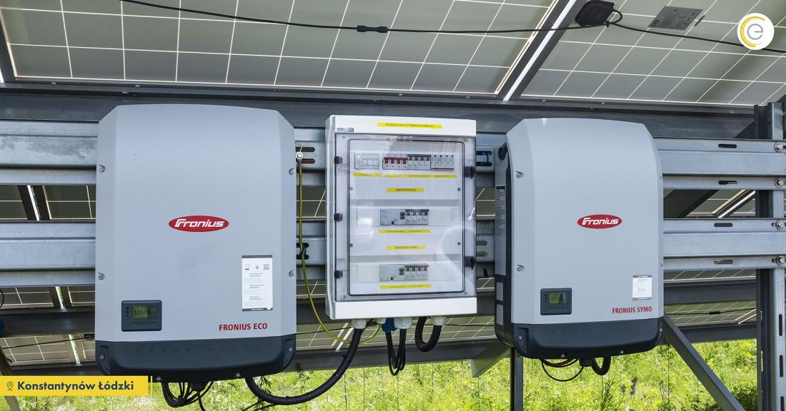 Dwa falowniki Fronius Eco i Fronius Symo zamontowane na metalowej konstrukcji pod panelami fotowoltaicznymi, z centralną skrzynką rozdzielczą z bezpiecznikami, widoczne okablowanie.