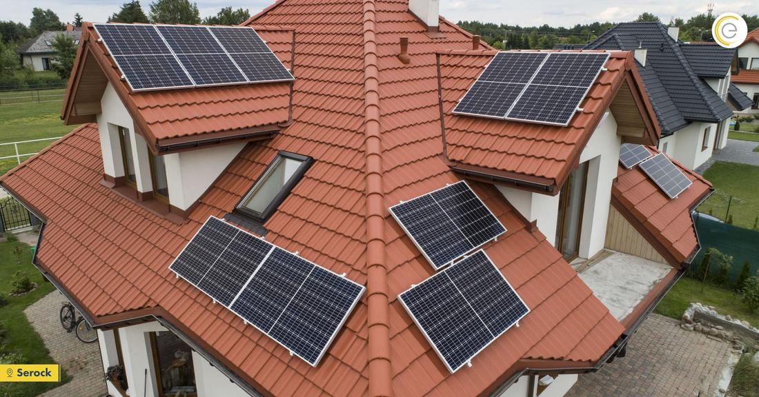 Widok z góry na dom z czerwoną dachówką i zamontowanymi panelami słonecznymi, w tle inne domy i zieleń.