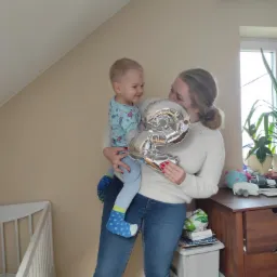 Uśmiechnięta kobieta trzymająca w ramionach małego chłopca, który patrzy na nią z uśmiechem, oboje trzymają srebrny balon w kształcie cyfry 2, w tle widoczne dziecięce łóżeczko i komoda z zabawkami.