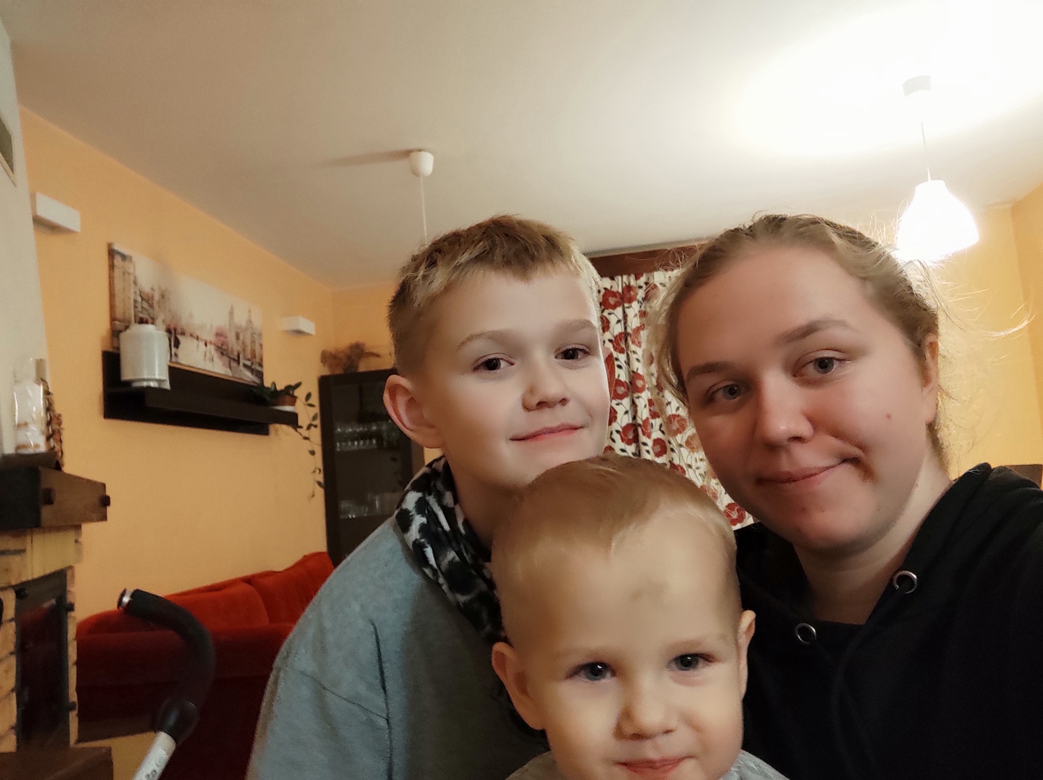 Portret kobiety z dwójką dzieci w domowym wnętrzu z beżowymi ścianami i czerwonym narożnikiem w tle.