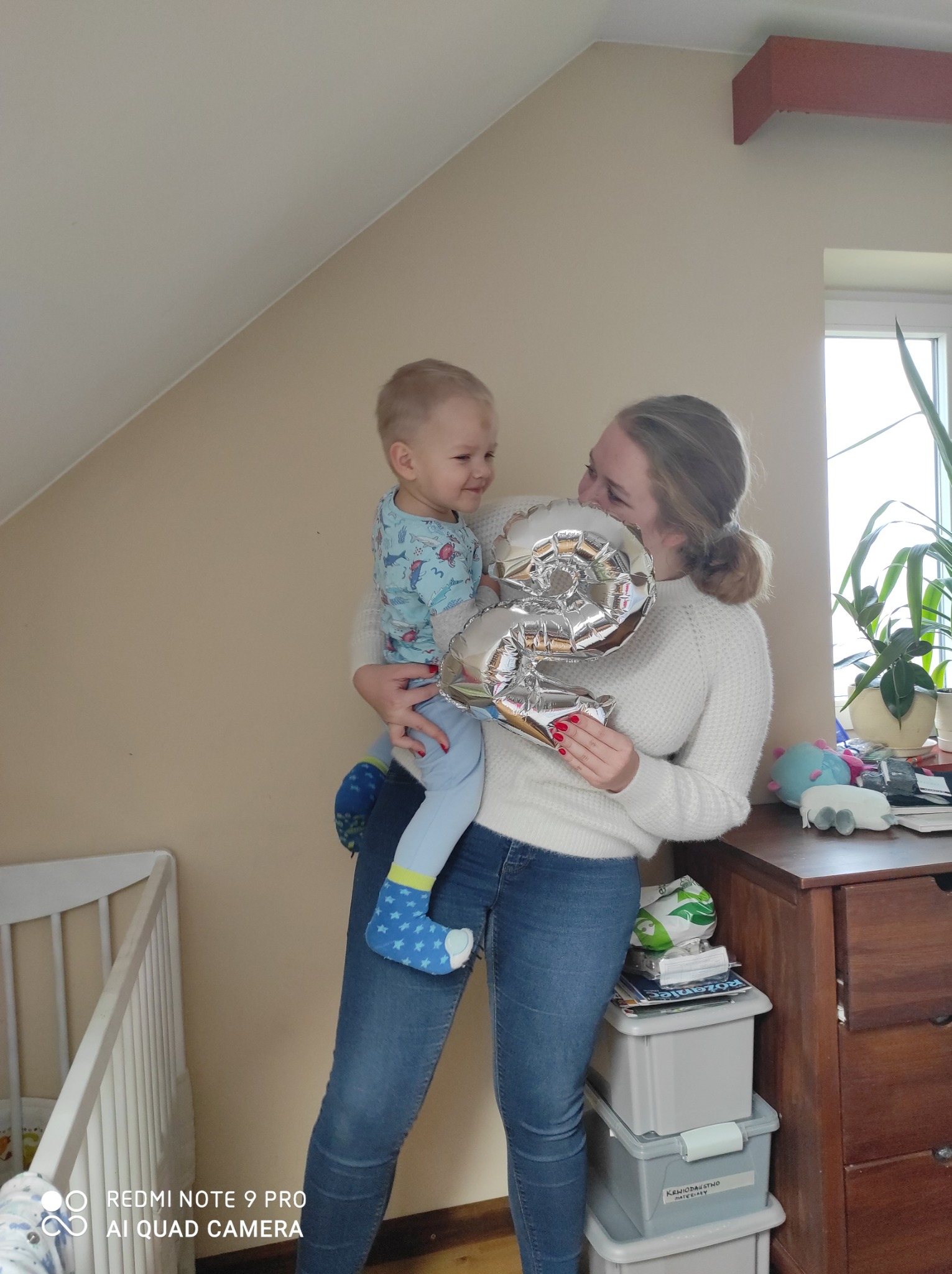 Uśmiechnięta kobieta trzymająca w ramionach małego chłopca, który patrzy na nią z uśmiechem, oboje trzymają srebrny balon w kształcie cyfry 2, w tle widoczne dziecięce łóżeczko i komoda z zabawkami.