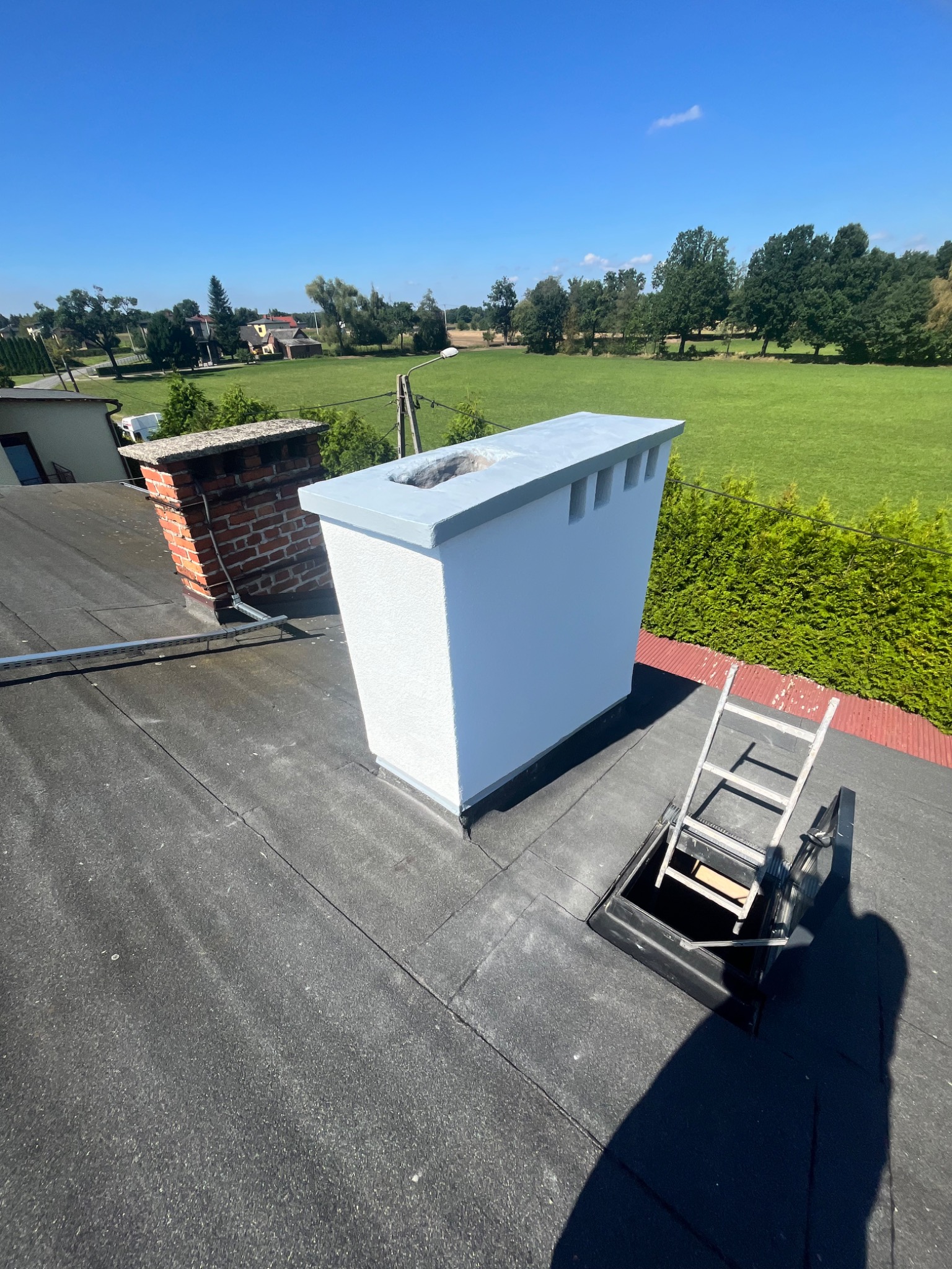 Biały, odnowiony komin wentylacyjny na czarnym dachu, obok ceglany komin. W tle zieleń drzew i błękitne niebo. Widoczny właz dachowy i drabina.
