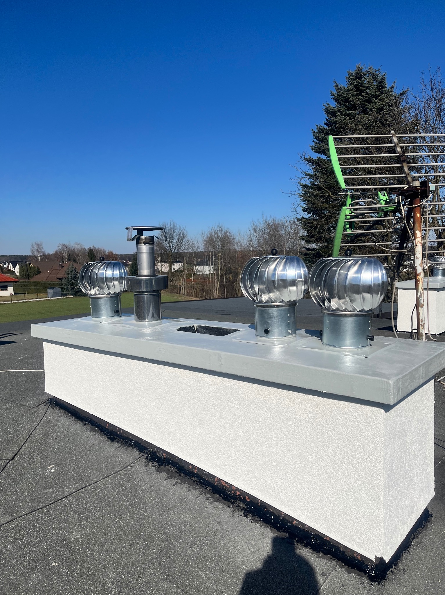 Cztery kominki wentylacyjne na płaskim dachu budynku, z widocznym niebieskim niebem i drzewami w tle. Jeden z kominków jest inny niż pozostałe.
