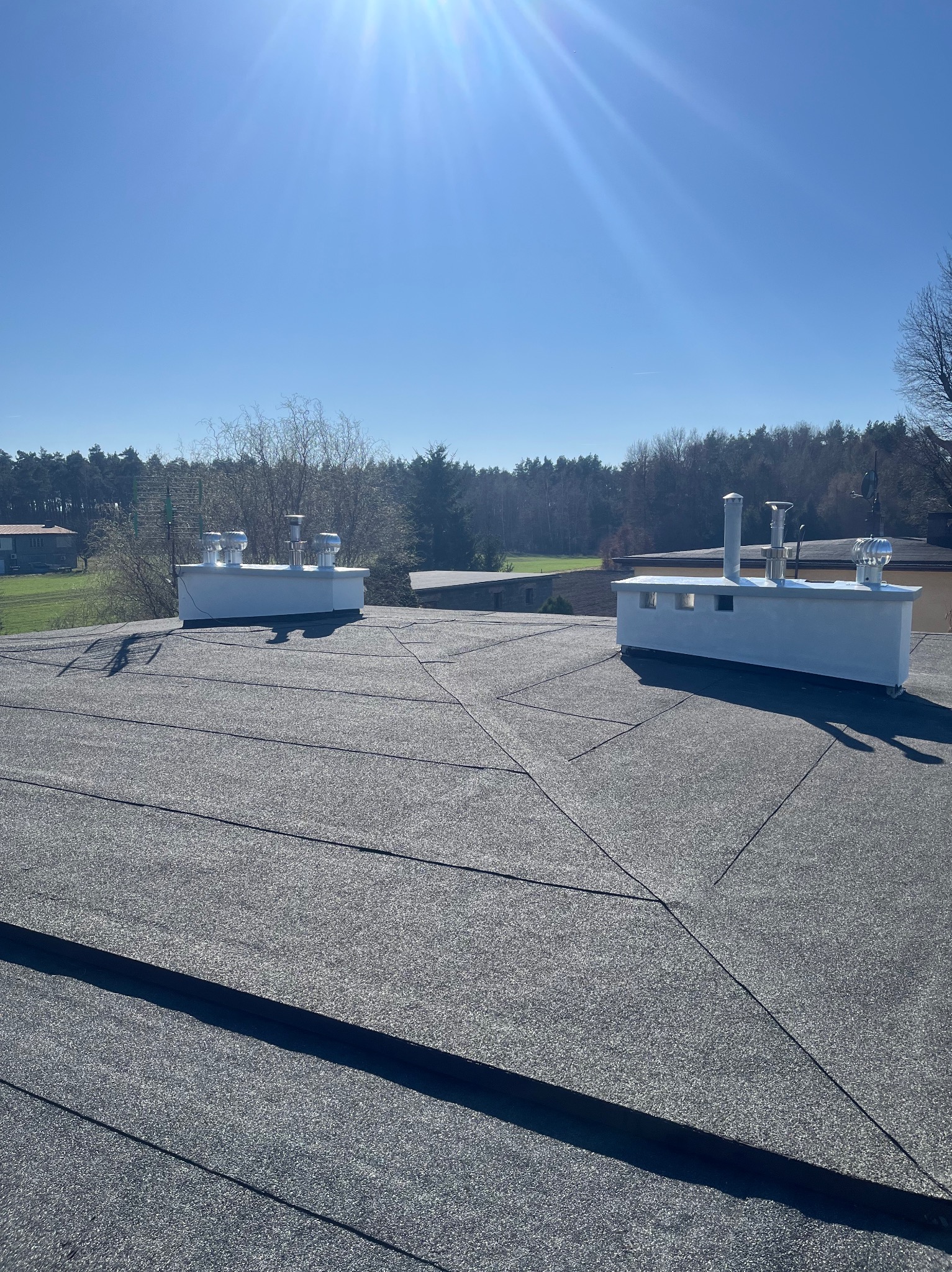 Czarny dach pokryty papą z dwoma białymi kominami wentylacyjnymi na tle zieleni i błękitnego nieba w słoneczny dzień. Widoczne łączenia papy.