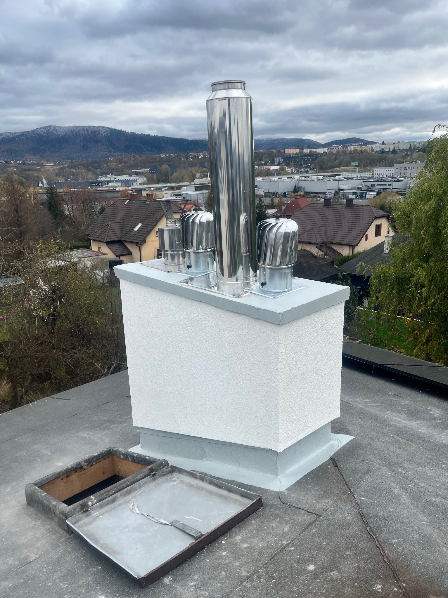 Biały, otynkowany komin z czterema metalowymi wylotami na dachu pokrytym papą. W tle zabudowa miejska i zalesione wzgórza pod pochmurnym niebem. Otwarty właz dachowy.