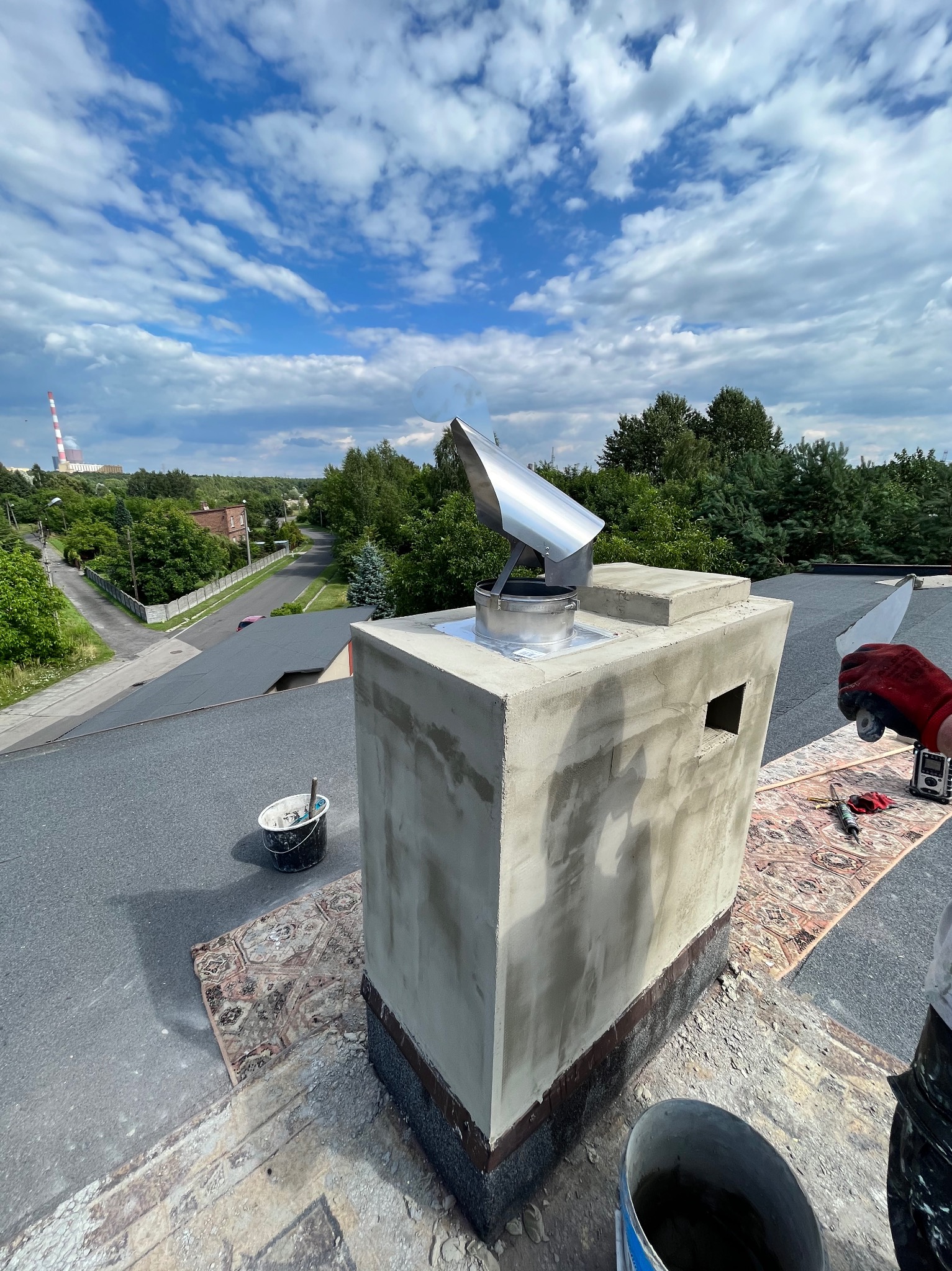 Wykończony komin z wkładem w Pszczynie, widok z góry. Na dachu leży dywan, obok stoją wiadra z zaprawą. W tle budynki i drzewa.