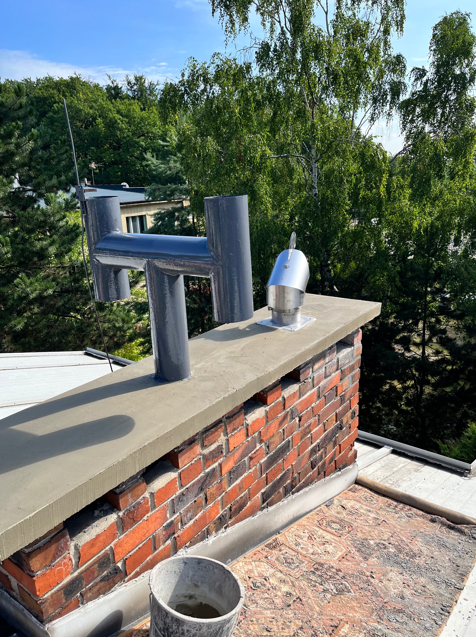 Odnowiony komin z cegły z nową obudową i metalowym wylotem, widok z góry na tle zieleni. Widoczny kubeł z zaprawą.