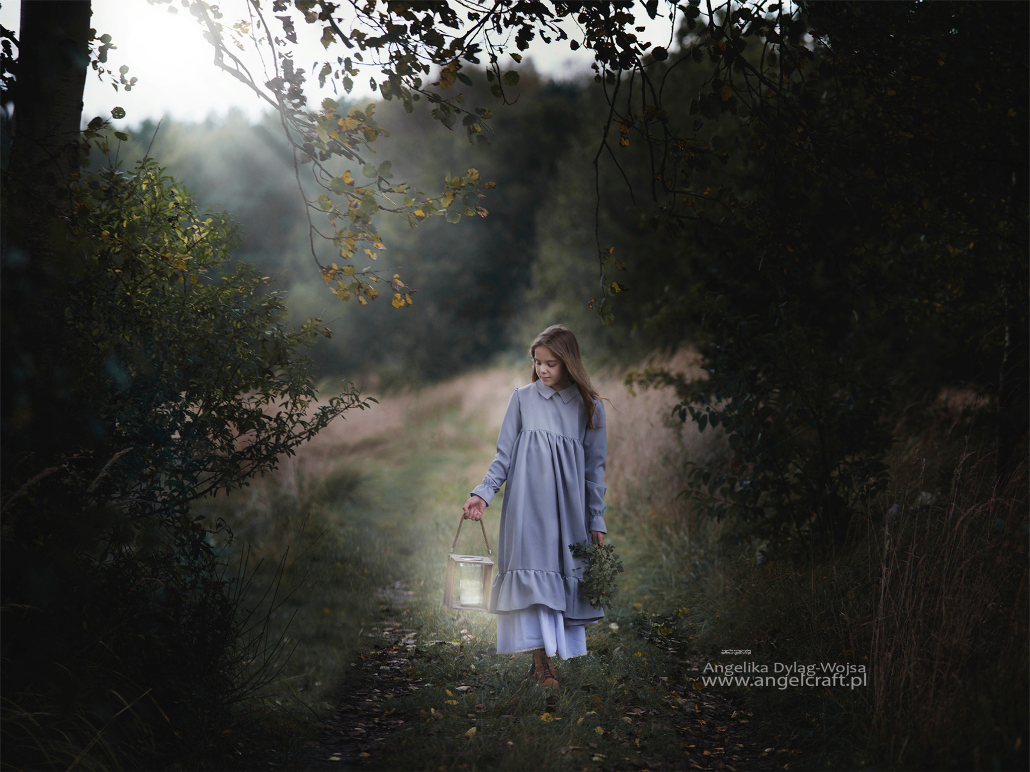 Dziewczynka w szarej sukience idzie leśną ścieżką, niosąc lampion i bukiet ziół, delikatne światło słoneczne przebija się przez korony drzew.