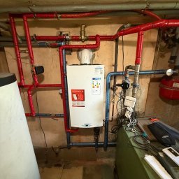 Lubas Serwis - Kocioł gazowy Ariston z instalacją: czerwone rury z izolacją, niebieskie rury wodne. Widoczne elementy armatury, zawory i przewody. W tle ściana i fragment zbiornika.