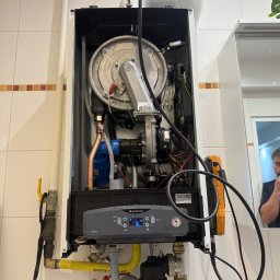 Lubas Serwis - Zamontowany, otwarty piec gazowy Ariston Cares S na ścianie z kafelkami. Widoczne rury, zawory, przewody, elementy sterujące i odbicie osoby w lustrze.