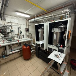 Lubas Serwis - Dwa nowoczesne piece gazowe, z widoczną instalacją hydrauliczną i rurami. Na pierwszym planie drabina z dokumentami i narzędziami. Całość w pomieszczeniu z beżowymi płytkami.