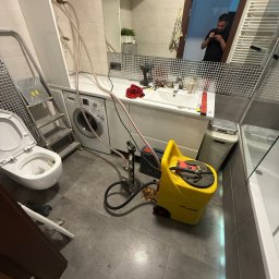 Lubas Serwis - Łazienka w trakcie prac hydraulicznych: podłączona toaleta, widoczna pralka, drabina, żółty sprzęt do testowania szczelności instalacji. W lustrze odbija się sylwetka osoby.