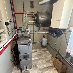 Lubas Serwis - Zmontowana instalacja grzewcza z piecem Defro, zbiornikiem akumulacyjnym i osprzętem. Widoczne rury, zawory i elementy sterujące. Podłoga wyłożona panelami imitującymi drewno.