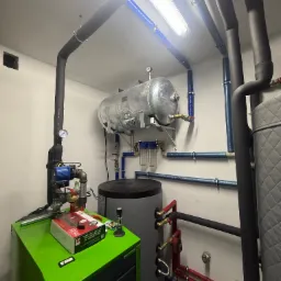 Zrealizowana instalacja grzewcza z zielonym piecem, zbiornikiem wyrównawczym i izolowanymi rurami w pomieszczeniu kotłowni z oświetleniem LED.