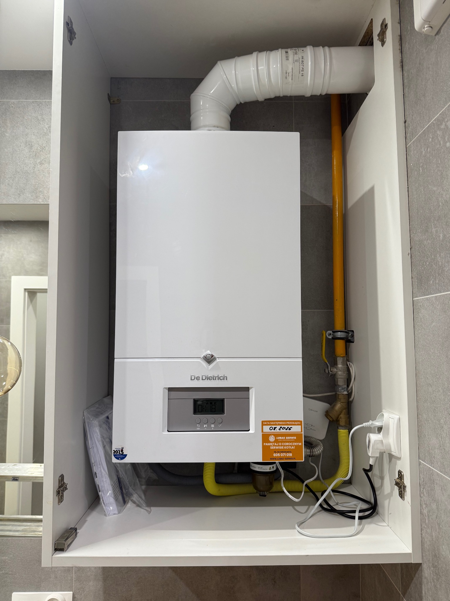 Biały piec gazowy marki De Dietrich zamontowany w szafce, z widocznymi rurami doprowadzającymi gaz i przewodem spalinowym. Na piecu naklejka informująca o dacie przeglądu.