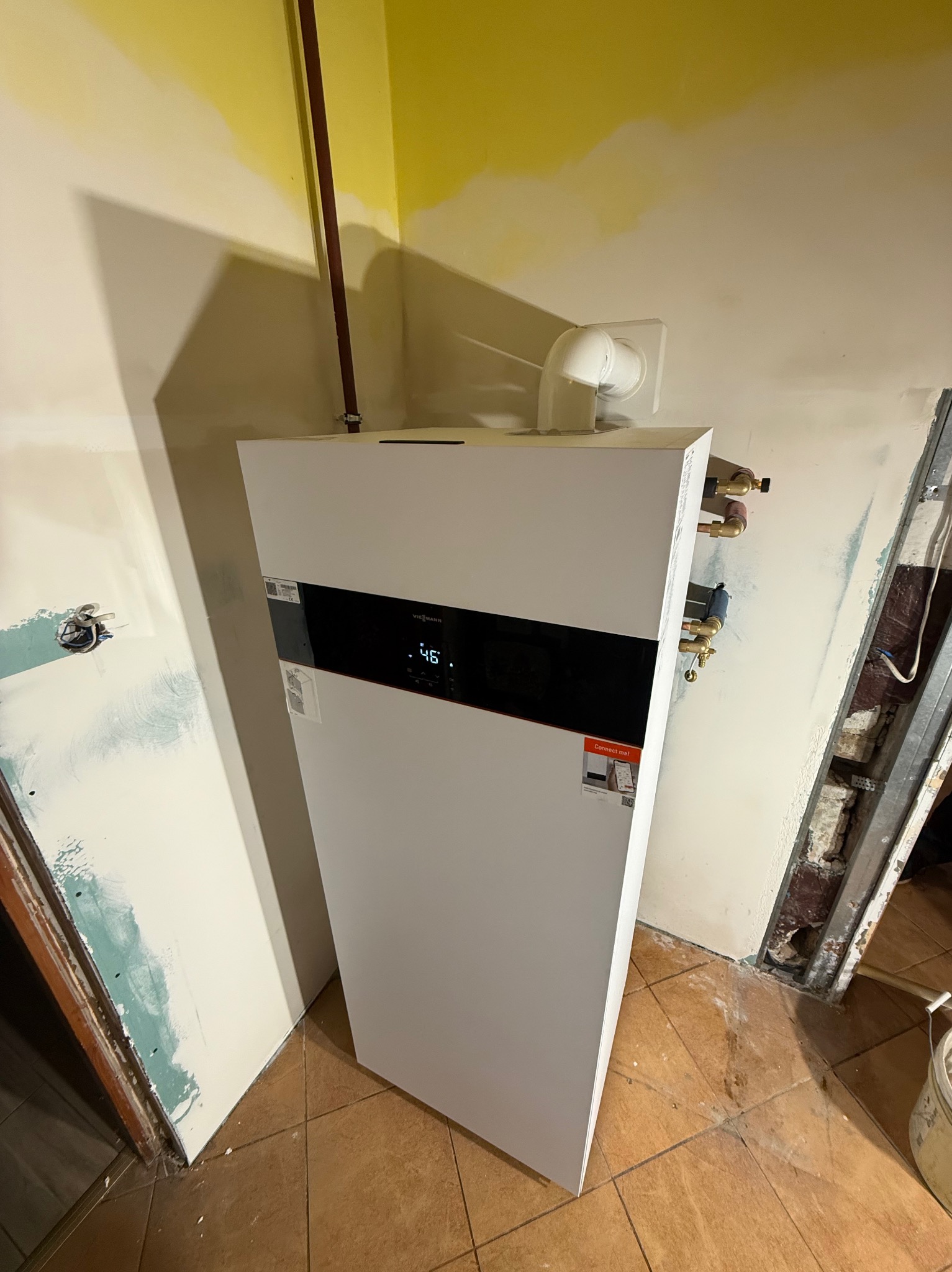 Biały piec gazowy Viessmann z czarnym panelem, zamontowany w pomieszczeniu w trakcie remontu, widoczne rury i elementy instalacji, brązowe płytki na podłodze.