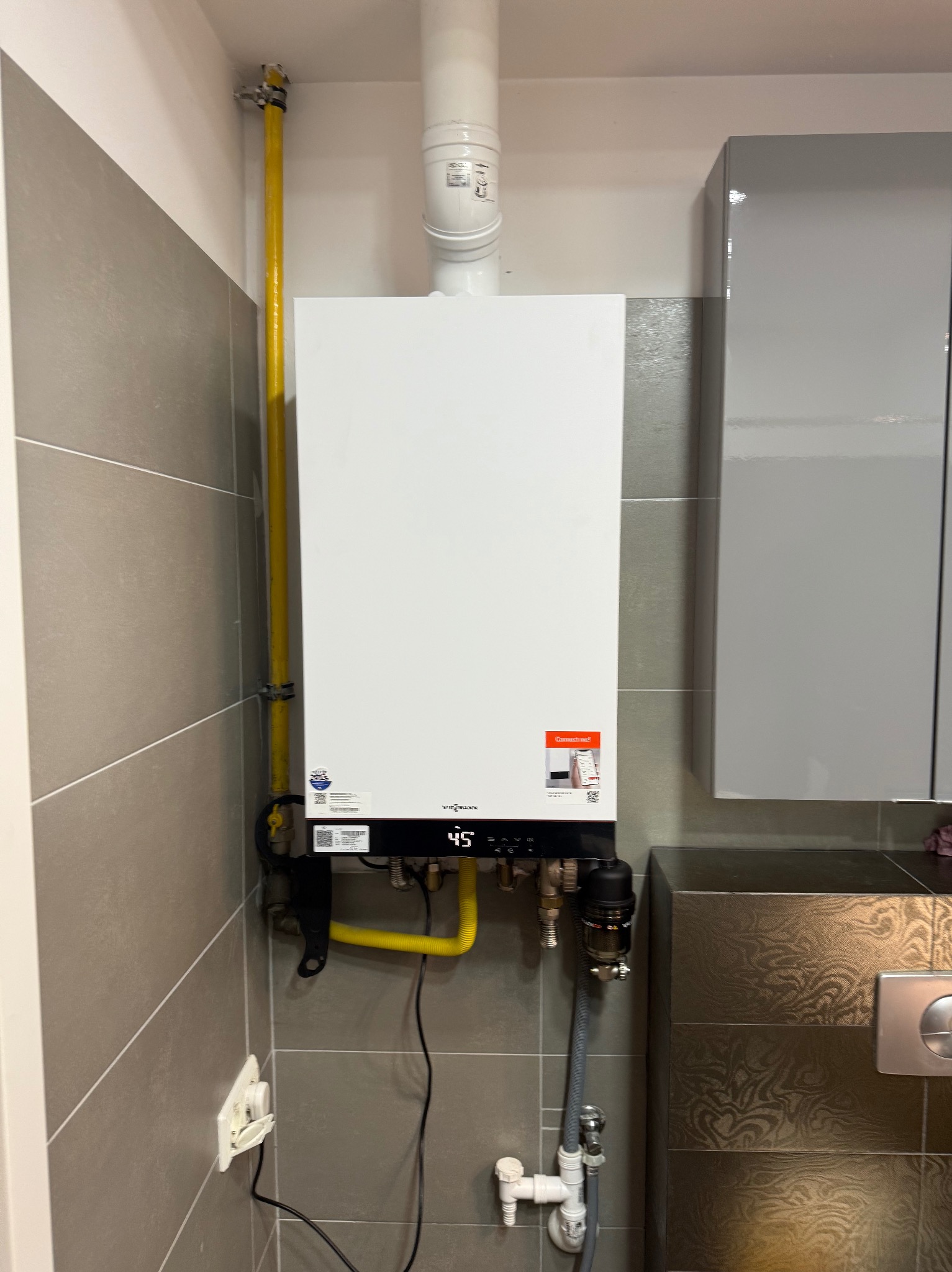 Biały piec gazowy Viessmann z żółtą instalacją, zamontowany na szarej, kafelkowanej ścianie w łazience, z widocznymi elementami instalacji i szafką w tle.