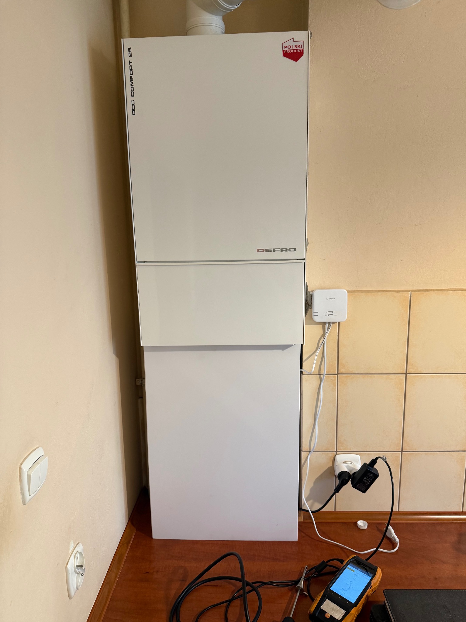 Biały, kompaktowy piec gazowy Defro z oznaczeniem 'Polski Produkt' na tle ściany z beżowymi płytkami. Na blacie widoczne narzędzia pomiarowe i kable.