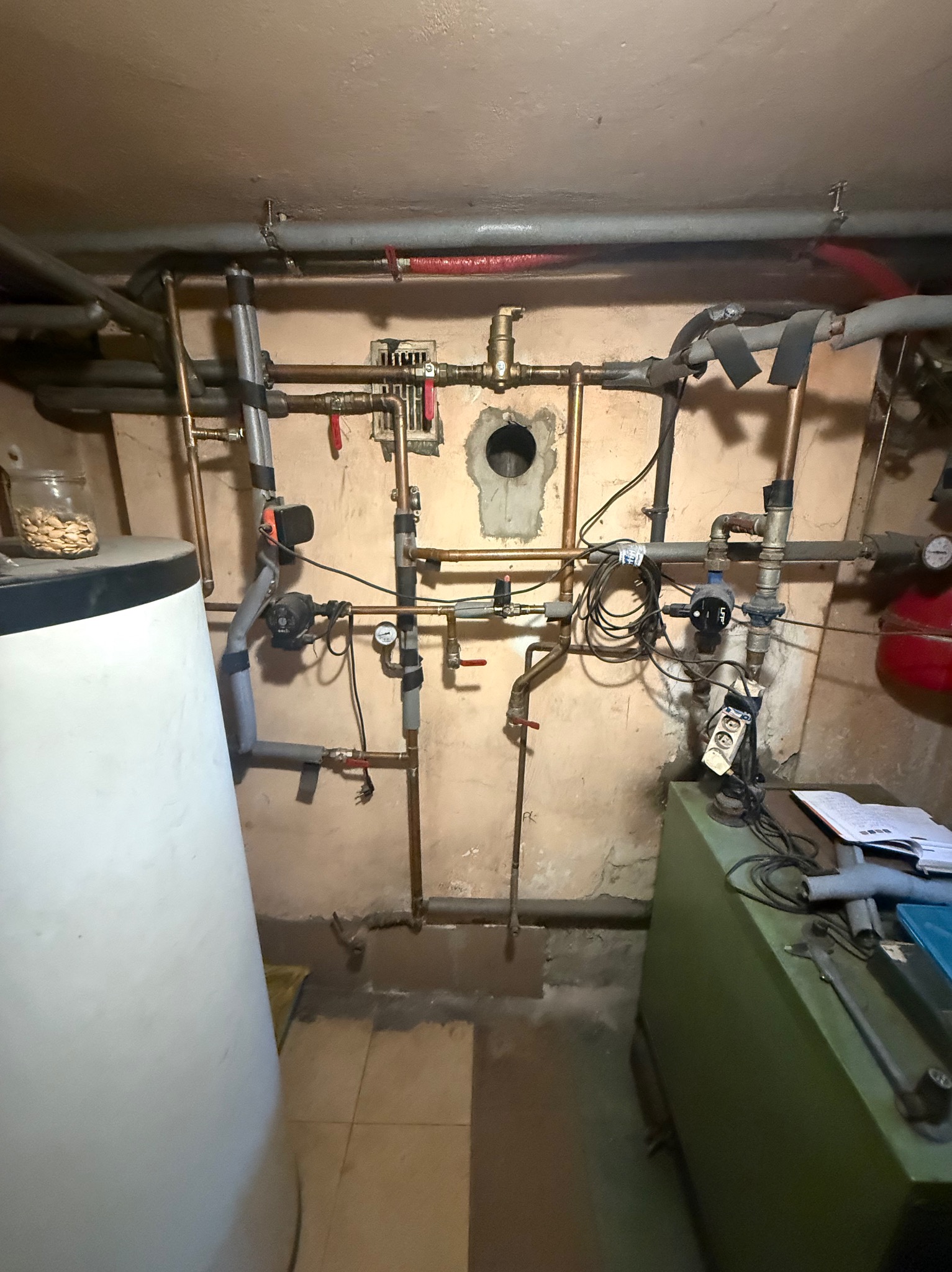 Złożona instalacja grzewcza z rurami miedzianymi, zaworami i pompami obiegowymi, zamontowana na ścianie w piwnicy. Widoczny zbiornik, manometry i izolacja rur.