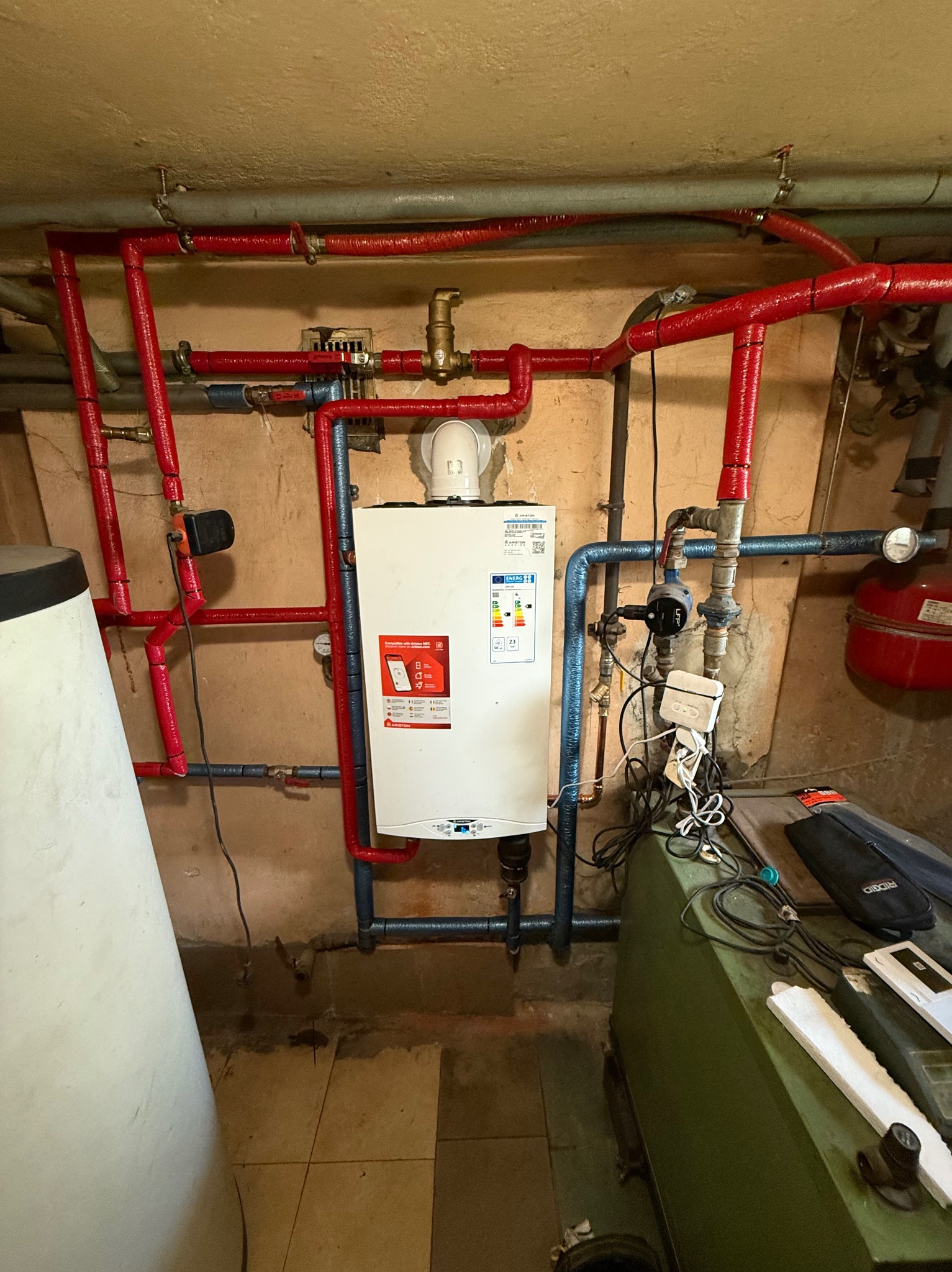 Kocioł gazowy Ariston z instalacją: czerwone rury z izolacją, niebieskie rury wodne. Widoczne elementy armatury, zawory i przewody. W tle ściana i fragment zbiornika.