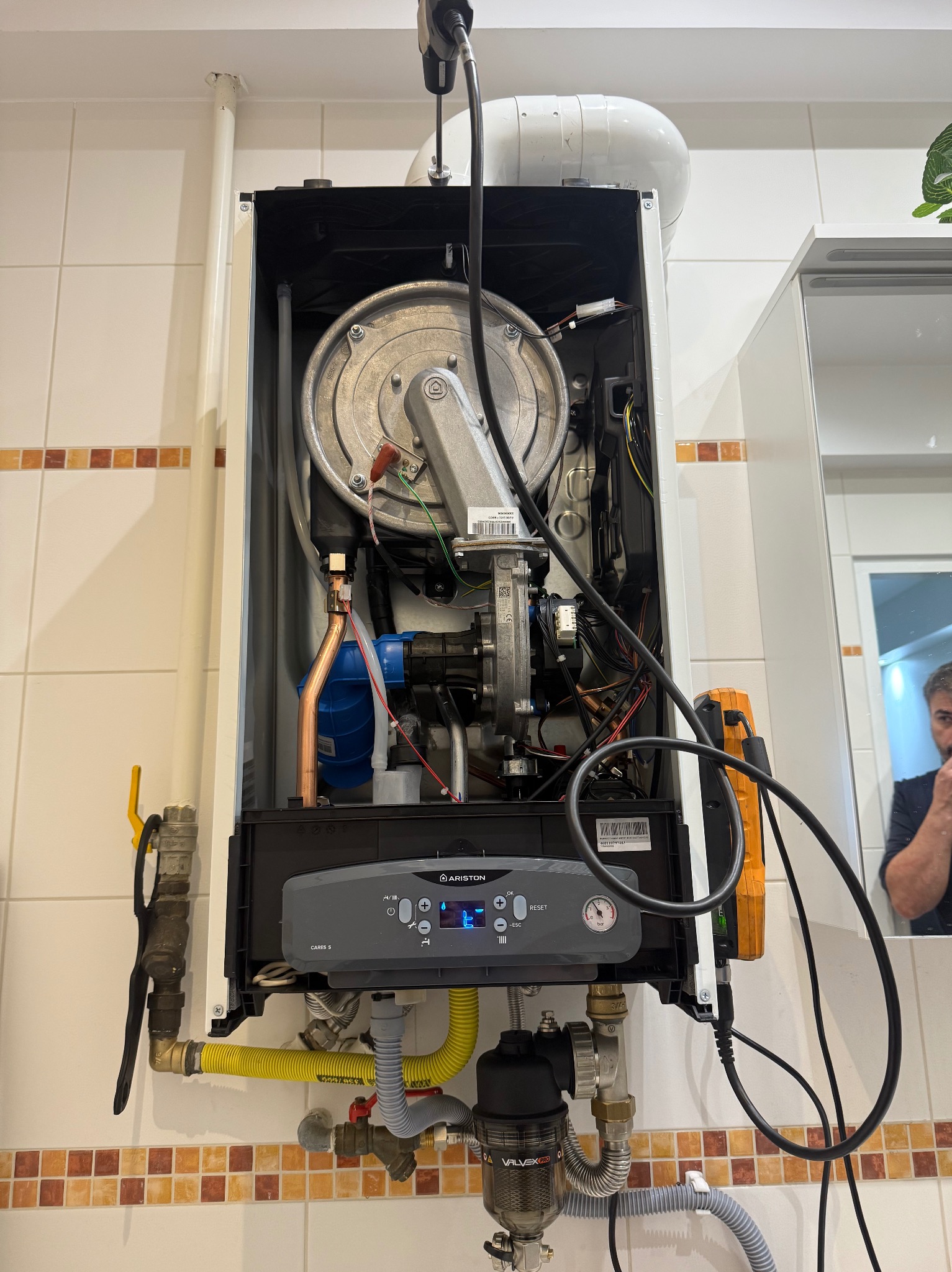 Zamontowany, otwarty piec gazowy Ariston Cares S na ścianie z kafelkami. Widoczne rury, zawory, przewody, elementy sterujące i odbicie osoby w lustrze.