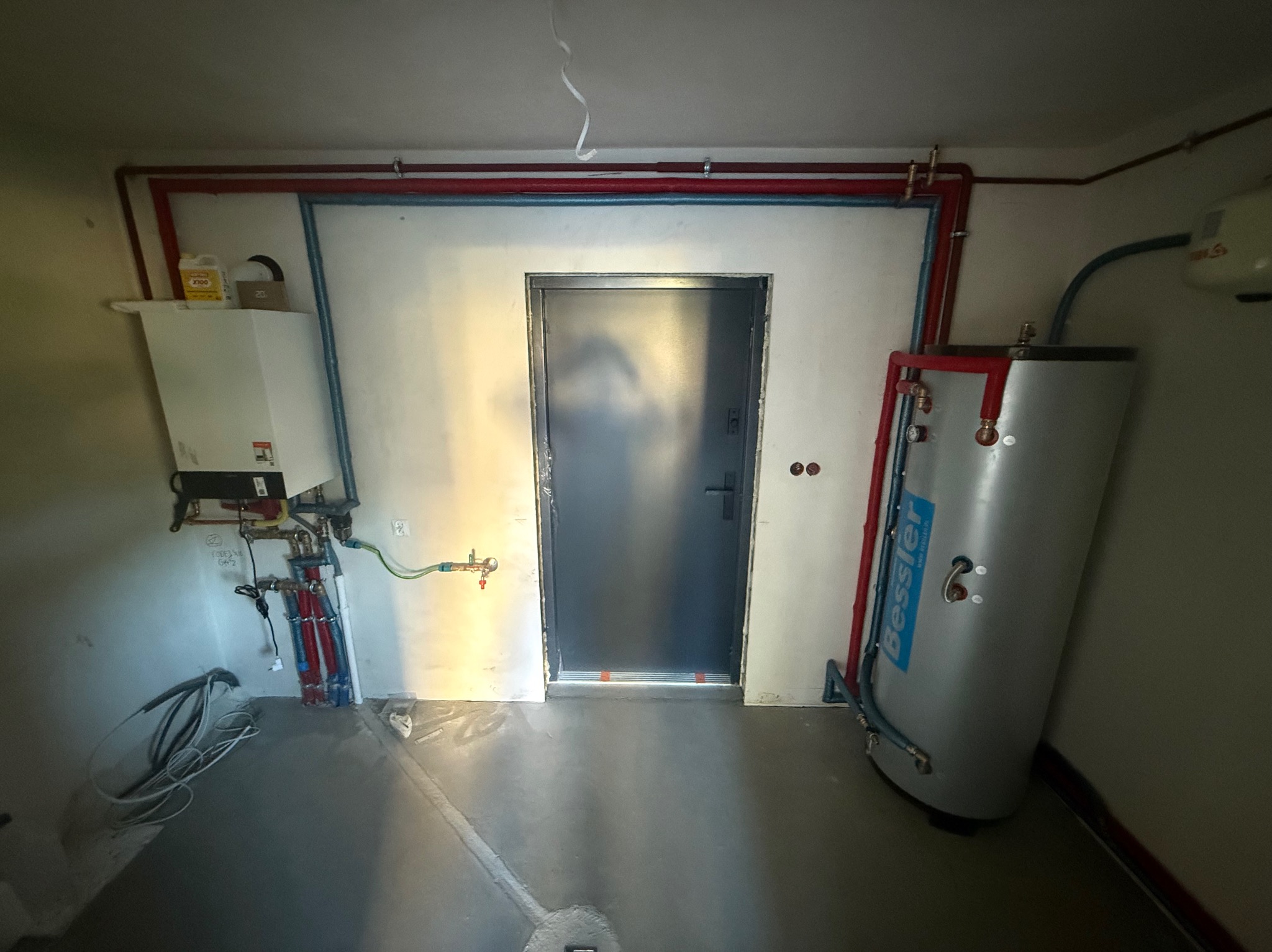 Kotłownia z piecem gazowym, zasobnikiem ciepłej wody i instalacją hydrauliczną. Widoczne rury w kolorze czerwonym i niebieskim. Ściany białe.