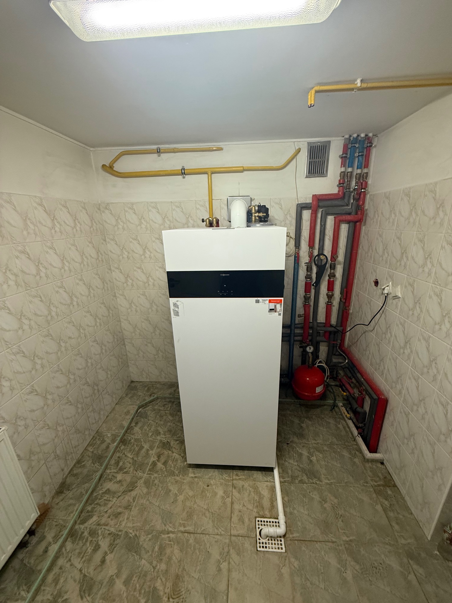 Biały piec gazowy Viessmann z podłączeniami hydraulicznymi i gazowymi w kotłowni wykończonej płytkami. Widoczne rury w kolorze czerwonym i szarym.