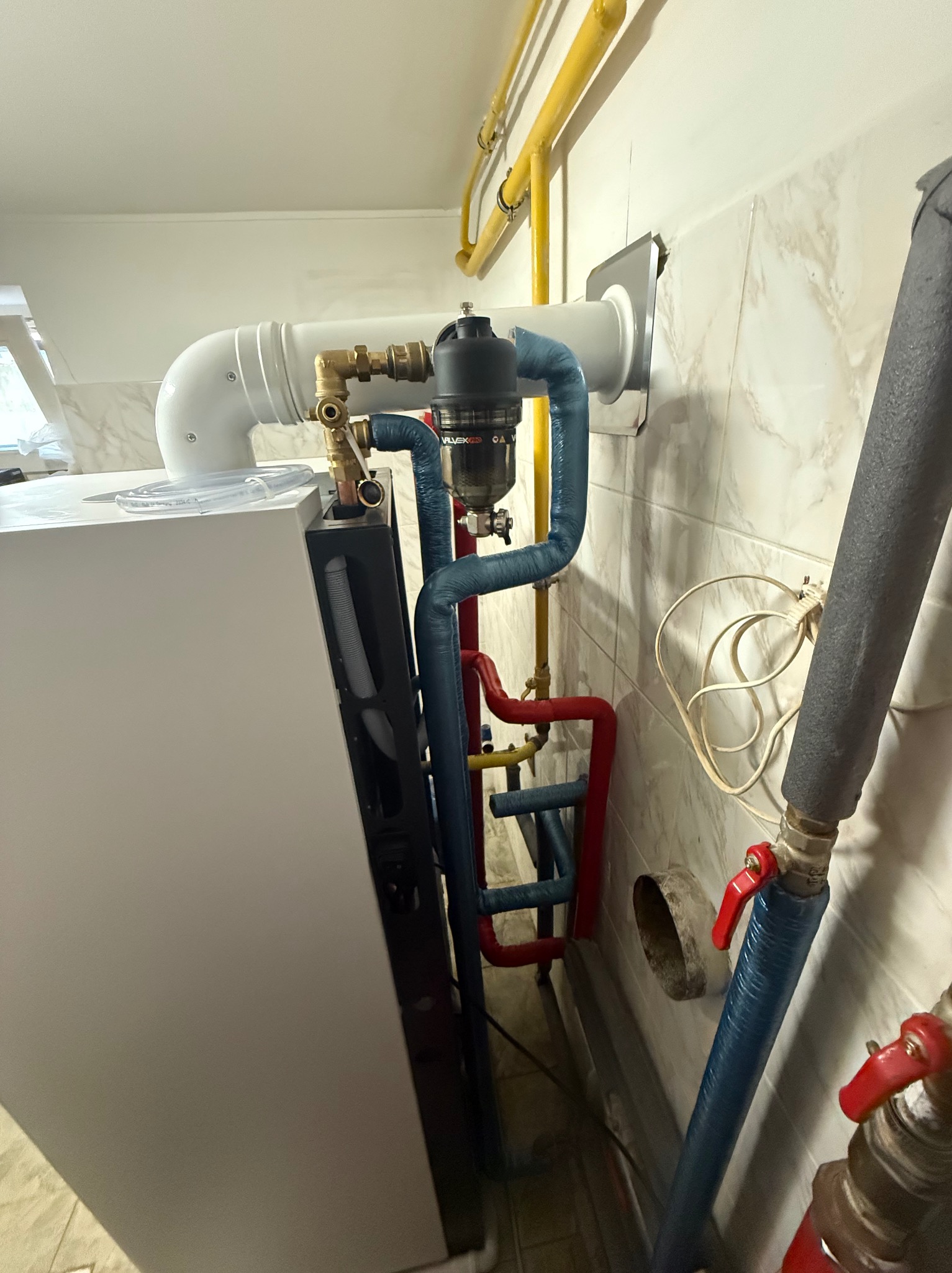 Nowoczesny piec gazowy z instalacją: niebieskie, czerwone i żółte rury. Widoczny filtr Valvex i biała obudowa pieca. Montaż w pomieszczeniu z marmurkową glazurą.