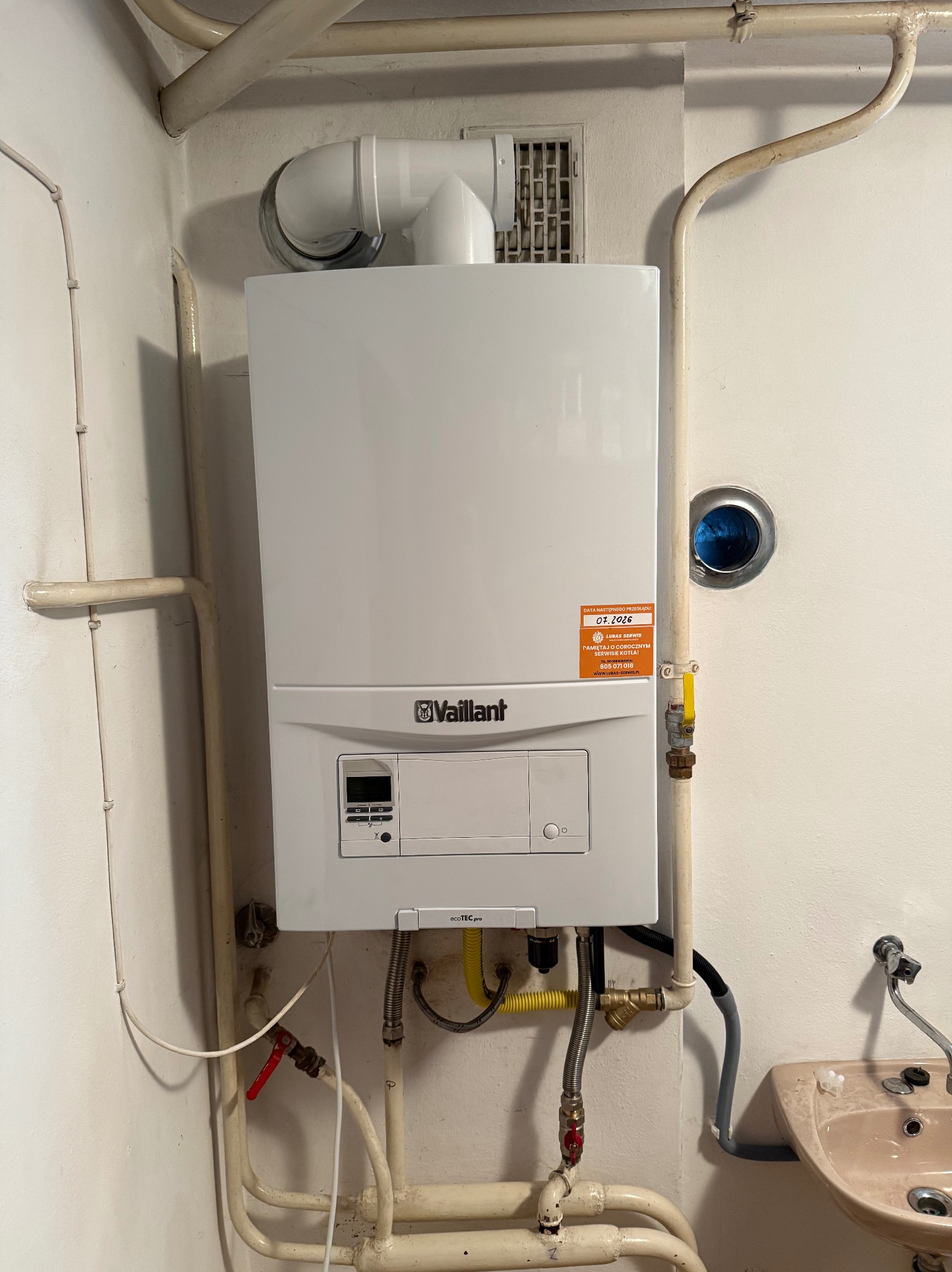 Biały, wiszący piec gazowy Vaillant z podłączeniami i naklejką serwisową w rogu pomieszczenia z rurami i umywalką. Widok z przodu, oświetlenie dzienne.